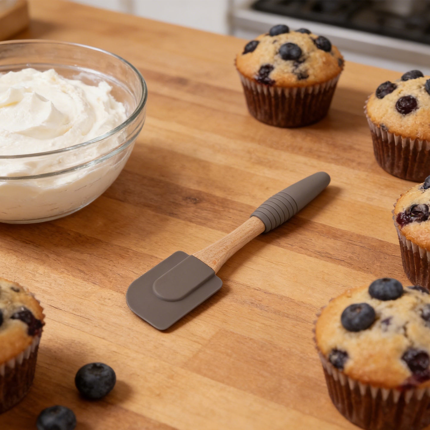 Blueberry muffins on a wooden table with a bowl of cream cheese frosting and a spatula.