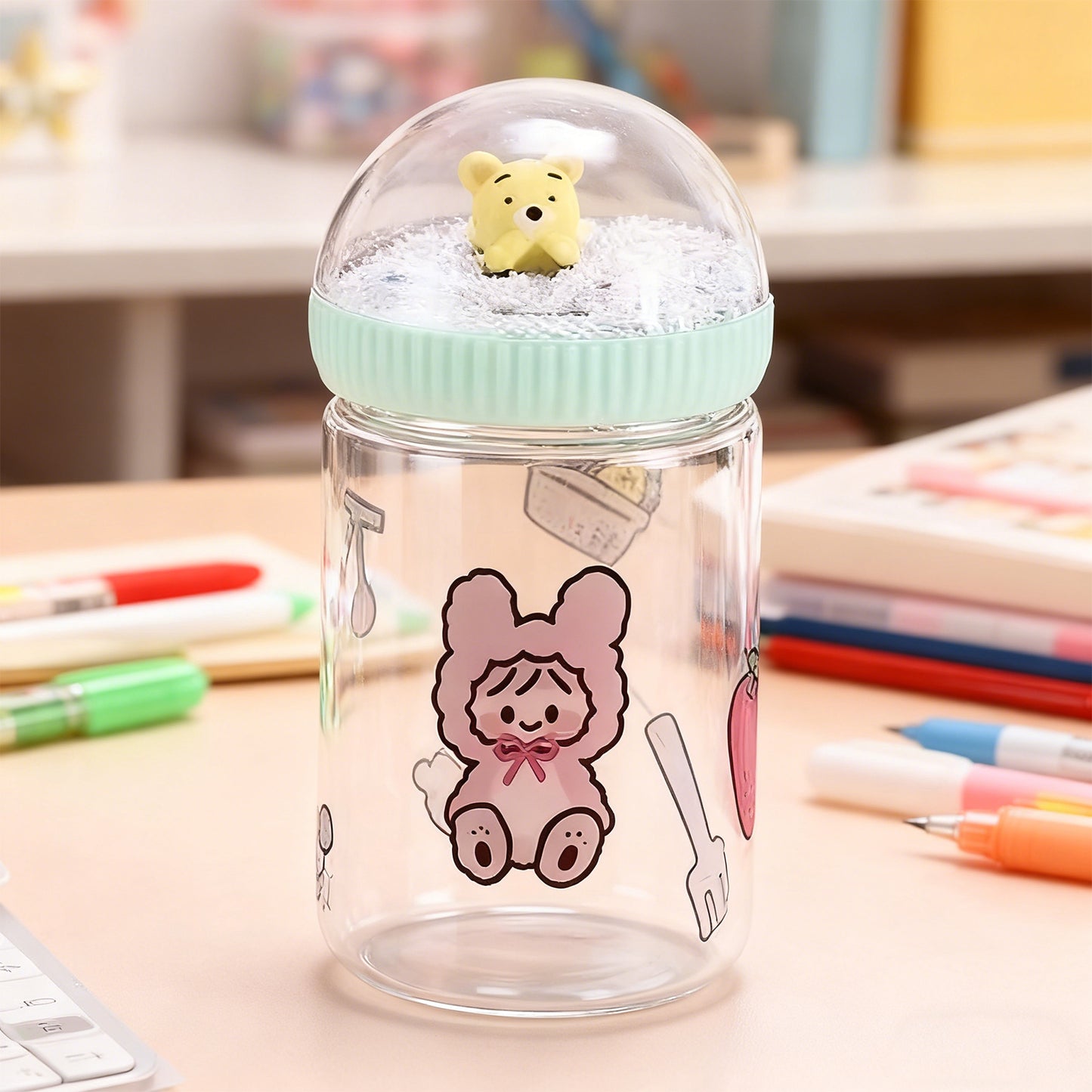 Clear jar with cartoon design and small yellow bear toy on a desk with stationery items.