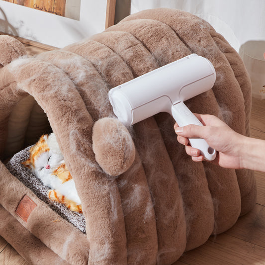 Person using a white pet grooming tool on a fluffy brown blanket with a cat underneath.